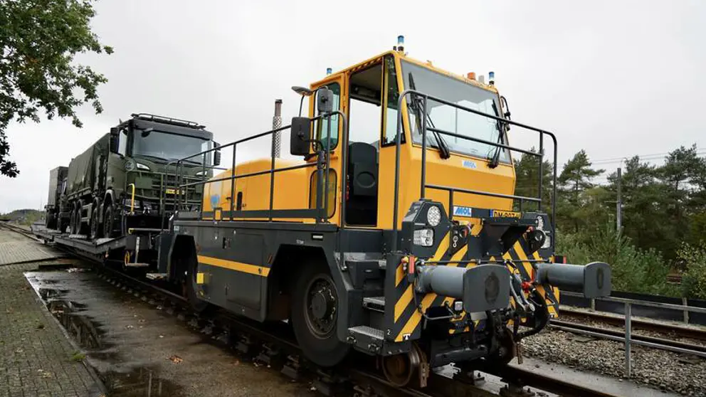 Een gele locomotief trekt groene Defensievrachtwagens.
