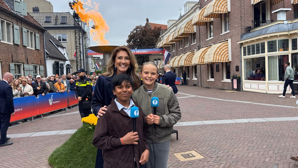 Minister Dilan Yeşilgöz met twee kindercorrespondenten met microfoons. Op de achtergrond het bevrijdingsvuur.