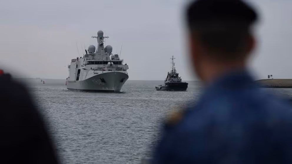 Het grijze schip De Vlissingen komt aangevaren. Rechts vaag een marinier in beeld.