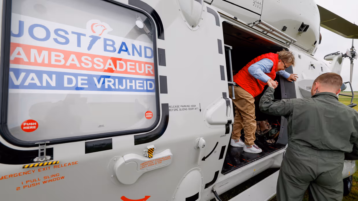 Een militair helpt een lid van de Jostiband uit de NH90 stappen. Op de helikopter staat de tekst ‘Jostiband, Ambassadeur van de Vrijheid’.