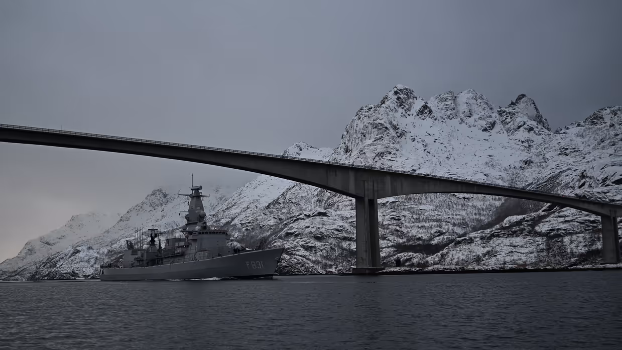Een militair schip vaart onder een brug door met bergen in de achtergrond.