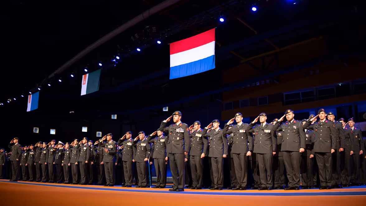 Militairen staan naast elkaar opgesteld terwijl ze salueren met boven hen de Nederlandse vlag.