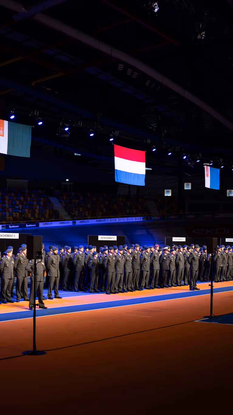 Militairen in dagelijks tenue staan naast elkaar opgesteld in een grote hal tijdens medaille-uitreiking.