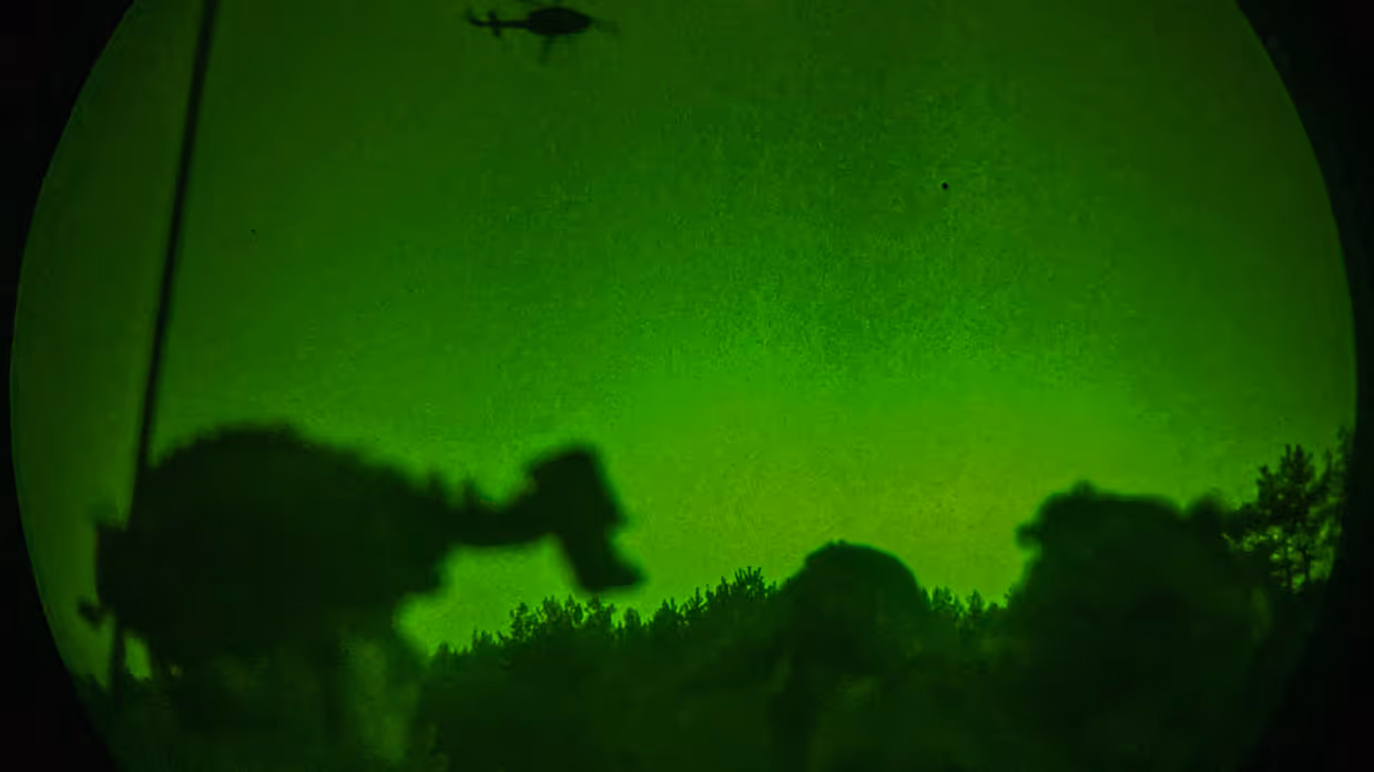 Een groep wachtende militairen zijn zichtbaar door een nachtlens, boven in de lucht vliegt een helikopter.