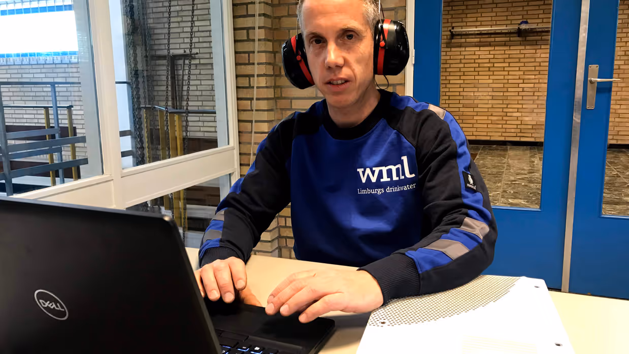 Jeroen Blomen met gehoorbescherming achter zijn laptop met een trui van Limburgse Waterleidingen aan. 