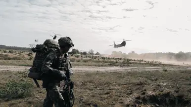 Een militair met zware bepakking verplaatst zich over de heide terwijl Chinook-helikopters laag overvliegen.