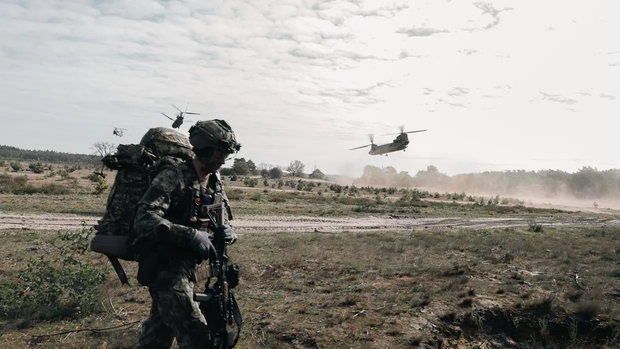 Een militair met zware bepakking verplaatst zich over de heide terwijl Chinook-helikopters laag overvliegen.