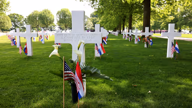 Een wit kruis met de tekst: Here rest in honored glory, a comrade in arms, known but to god. In de achtergrond nog meer kruisen, allemaal voorzien van een klein Amerikaans en Nederlands vlaggetje en een aantal is voorzien van bloemen.  