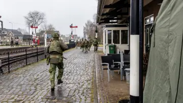  foto links: Militairen installeren een ronde satellietschotel, omringd door uitrusting en voertuigen. Links midden: Een groep soldaten loopt over een keienpad langs een station met een overdekt terras; een soldaat loopt voorop, de rest volgt in een rij. Rechts midden: Drie militairen werken op een open veld: één stelt een grote satellietschotel af, twee anderen bedienen apparatuur en kabels op de grond. Rechts: Vier militairen in gevechtstenue lopen over een grasveld langs een onverharde weg;