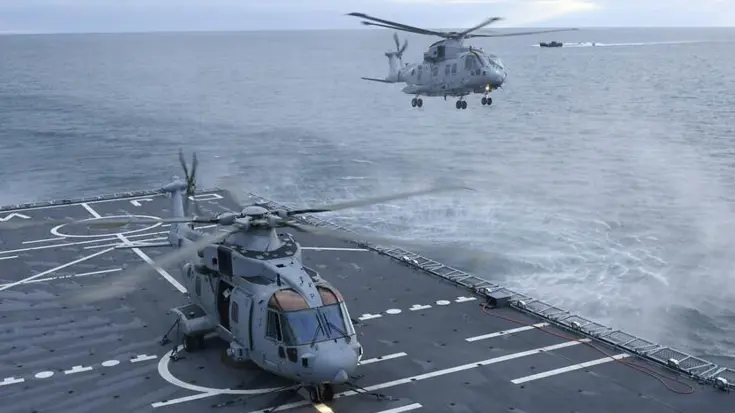 Een helikopter vliegt boven landingsdek schip. Een ander toestel staat op het dek.