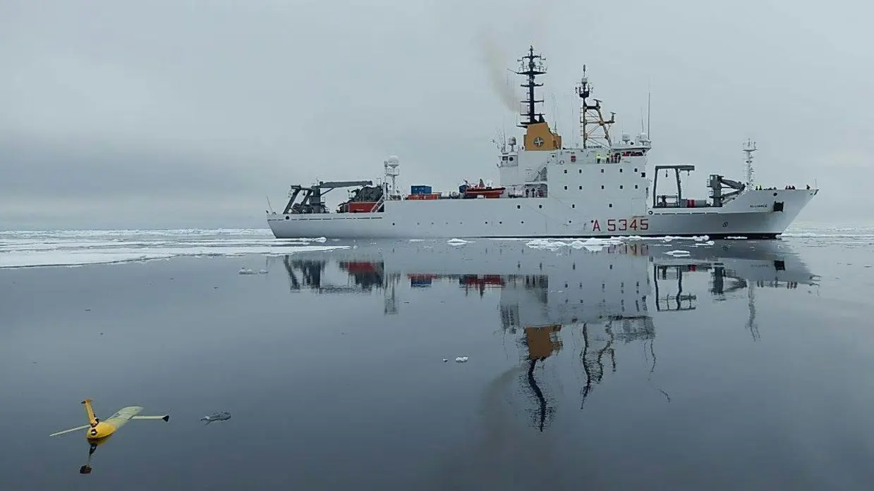 Een wit onderzoeksvaartuig op zee, met op de voorgrond een varende drone.