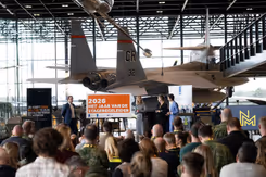 Publiek kijkt naar podium met mensen en een oranje bord. Daarachter hangt een gevechtsvliegtuig.