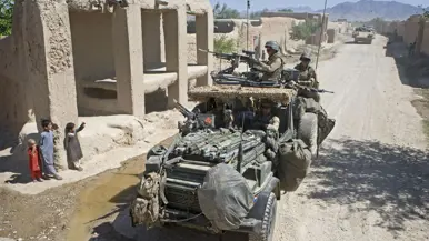 Alt-tekst: Een militair zit op een verhoging van zand en geeft een snoepje aan een meisje. Achter haar staat een jongen. 