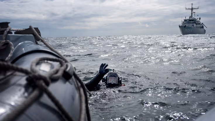 Een duiker in zee naast een marineschip.