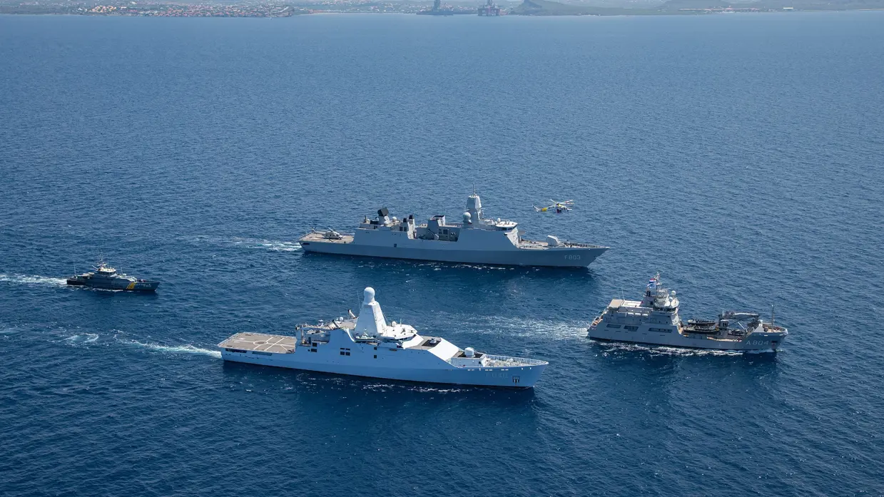 4 marinevaartuigen varen in formatie op open zee, met een helikopter erboven en land op de achtergrond.