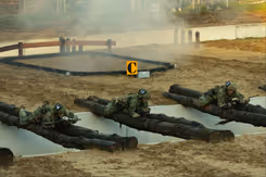 Een greppel met boomstammen die eroverheen liggen waar militairen overheen kruipen en rook oop  de achtergrond.