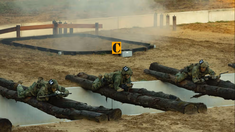 Een greppel met boomstammen die eroverheen liggen waar militairen overheen kruipen en rook oop  de achtergrond.