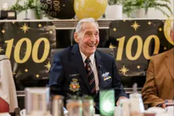 Ton Boelens, zittend aan een tafel, met de cijfers 100 aan beide zijden op de muur erachter.