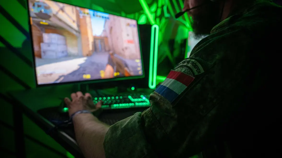 Een militair toont zijn geweer aan een burger. Een militair speelt een videogame.