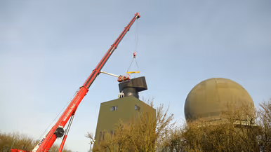 Links plaatst hijskraan een radar op een hoge toren. Rechts steekt een radarkoepel boven begroeiing uit.