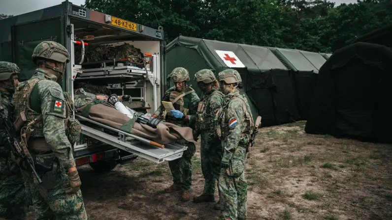Alttekst: 5 militairen buigen zich over een gewonde collega die wordt ingeladen in een militair voertuig. 