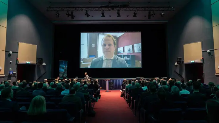 Staatssecretaris op groot scherm. Zaal mensen kijkt.