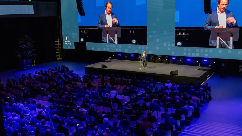 Staatssecretaris Tuinman staat op een podium. Achter hem is hij zichtbaar op 2 schermen. 