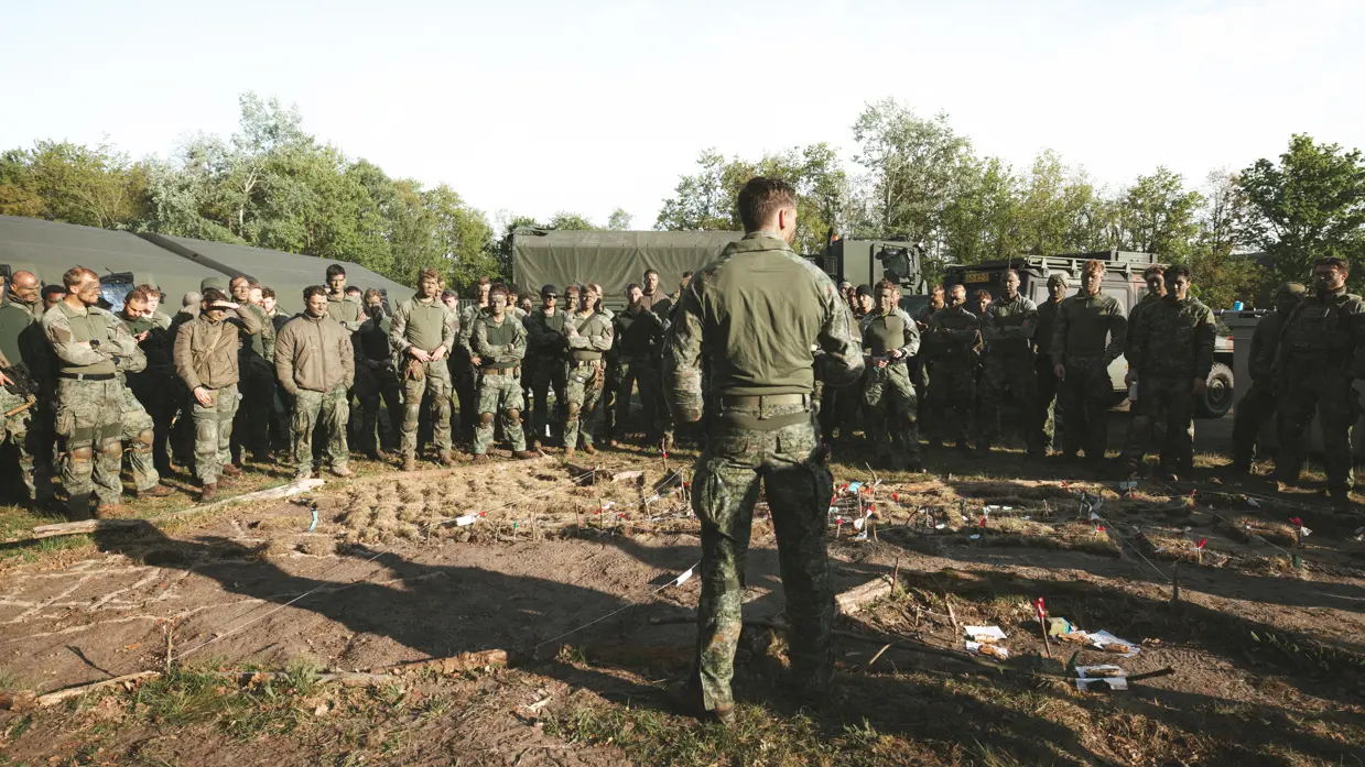Militairen verzamelen zich rond een zandmodel tijdens een briefing in het veld.