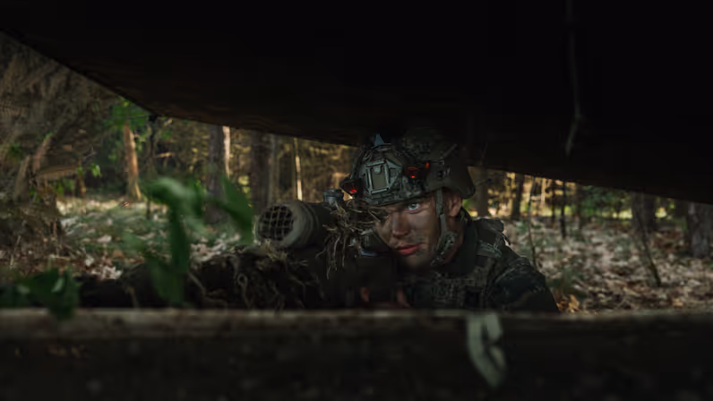 Een gecamoufleerde militair ligt met zijn wapen in stelling onder een schuilnet in een bosgebied tijdens oefening Versatile Guard. 