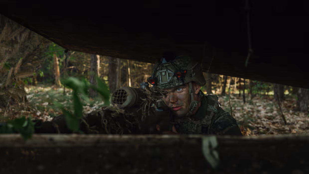 Een gecamoufleerde militair ligt met zijn wapen in stelling onder een schuilnet in een bosgebied tijdens oefening Versatile Guard. 