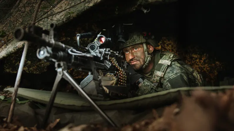 Een militair ligt met een machinegeweer in stelling onder een gecamoufleerde schuilpositie.
