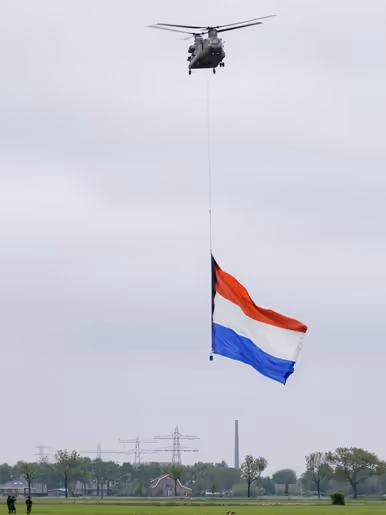 Een Chinook hangt stil boven een weiland met eronder een enorme Nederlandse vlag. Links onder staan drie mensen. Midden onderin staan een boerderij, een paar huizen, hoogspanningsmasten en een hoge schoorsteen.