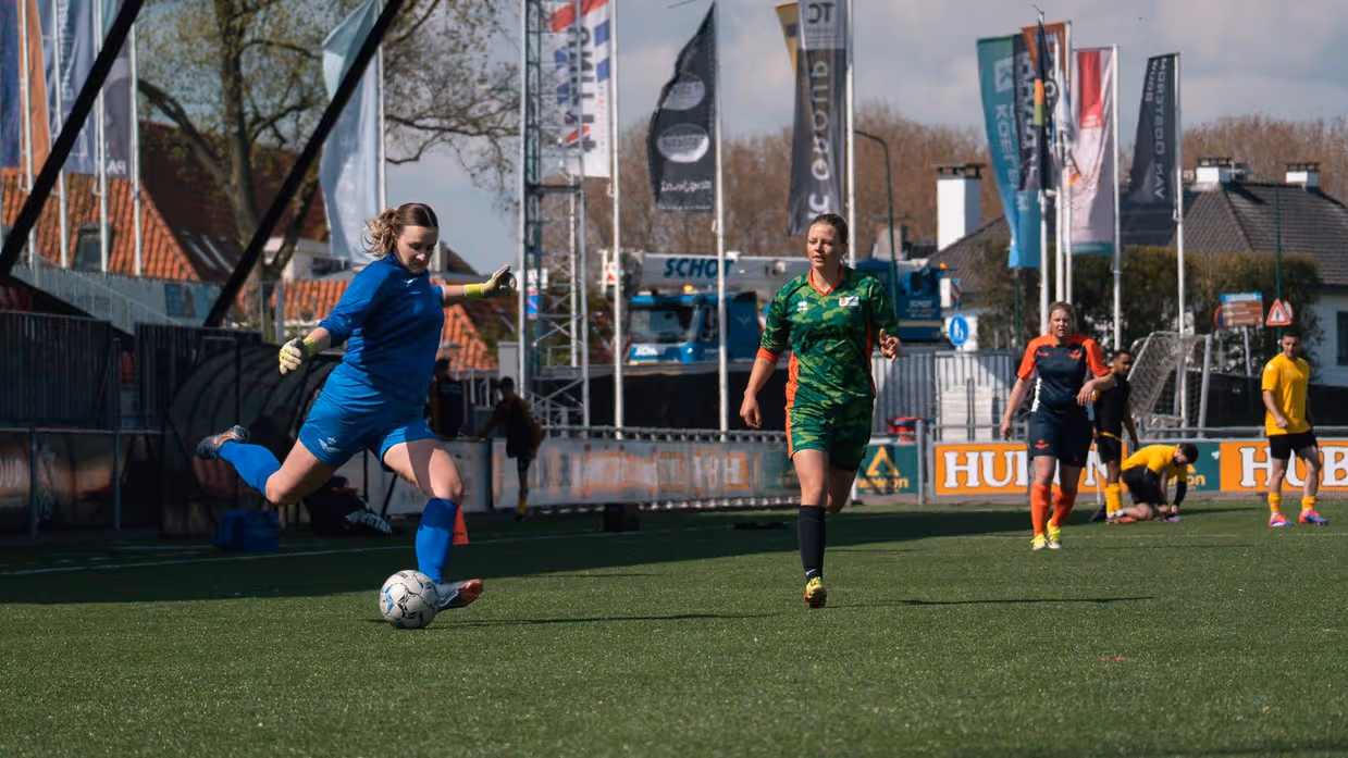 Op een voetbalveld met een tribune aan de zijkant schiet een vrouw in een blauw shirt de bal, terwijl andere speelsters in een groen shirt komen aanrennen. 