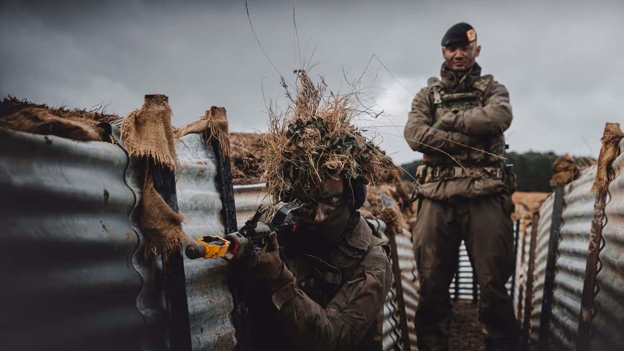 Een militair staat met zijn armen over elkaar. Hij kijkt mee over de schouders van een collega die oefent met een wapen in een loopgraaf.