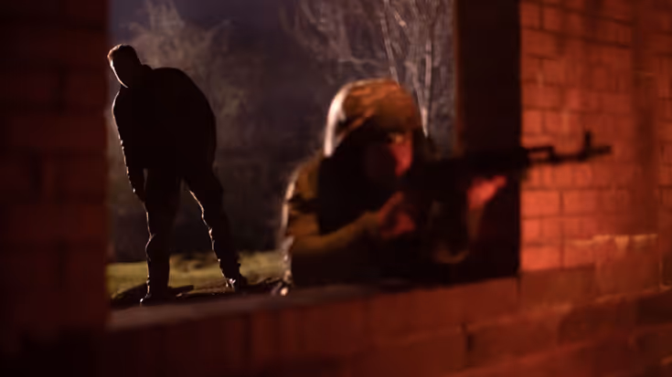 Militair leunt door raam en richt wapen. Een instructeur kijkt op de achtergrond mee.