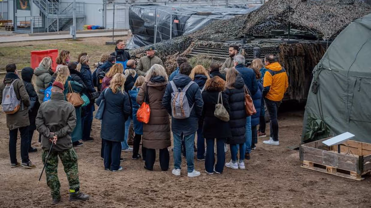 Mensen in burger luisteren naar militair voor de groep. Op de achtergrond militair materieel.