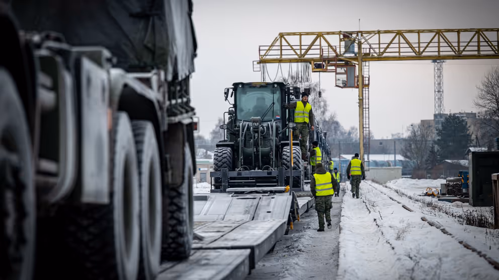 Mannen in gele hesjes lopen langs een treinstel waar militaire voertuigen op staan. 