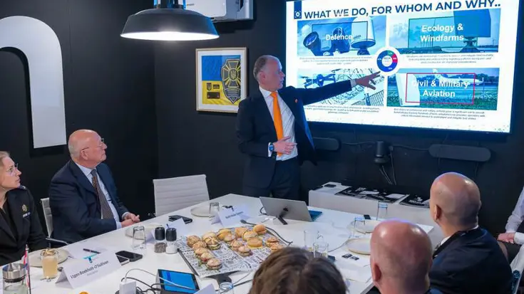 Tafel waar mensen aanzitten. Een staat en wijst naar groot scherm.