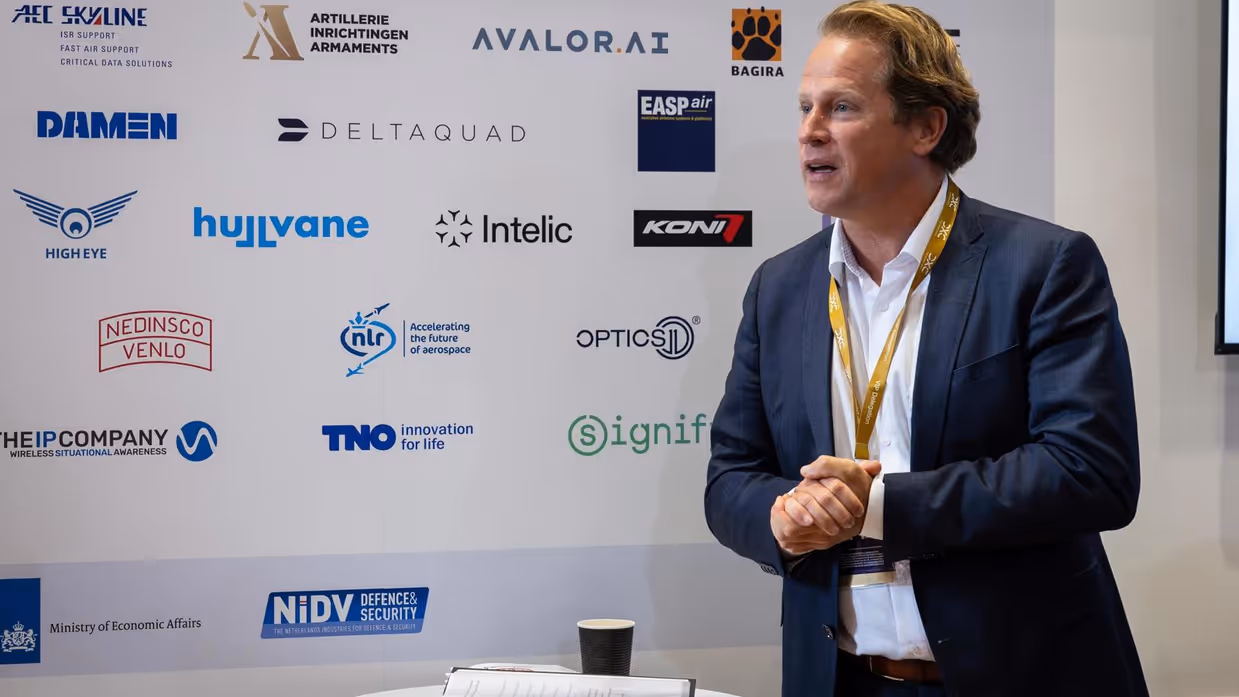 De staatssecretaris houdt een toespraak. Achter hem zijn de namen van een aantal bedrijven zichtbaar. 