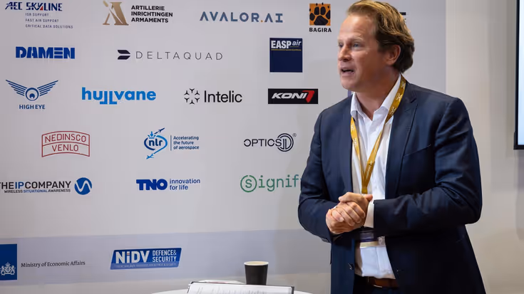 De staatssecretaris houdt een toespraak. Achter hem zijn de namen van een aantal bedrijven zichtbaar. 